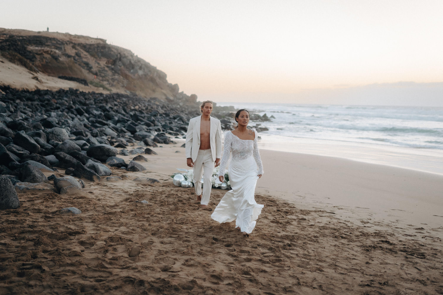Ein Brautpaar im Disko-Hochzeitsstil geht nach Sonnenuntergang Hand in Hand am Strand von Fuerteventura auf den Betrachter zu.