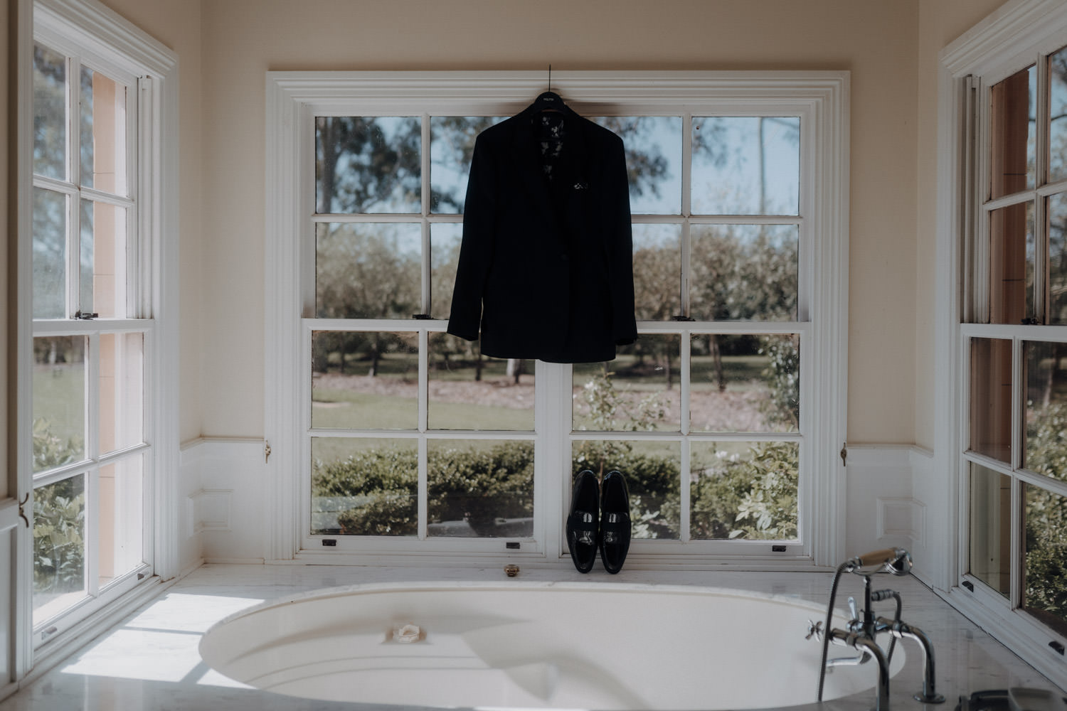 Das Jacket eines Bräutigams hängt im Badezimmer am Fensterrahmen.