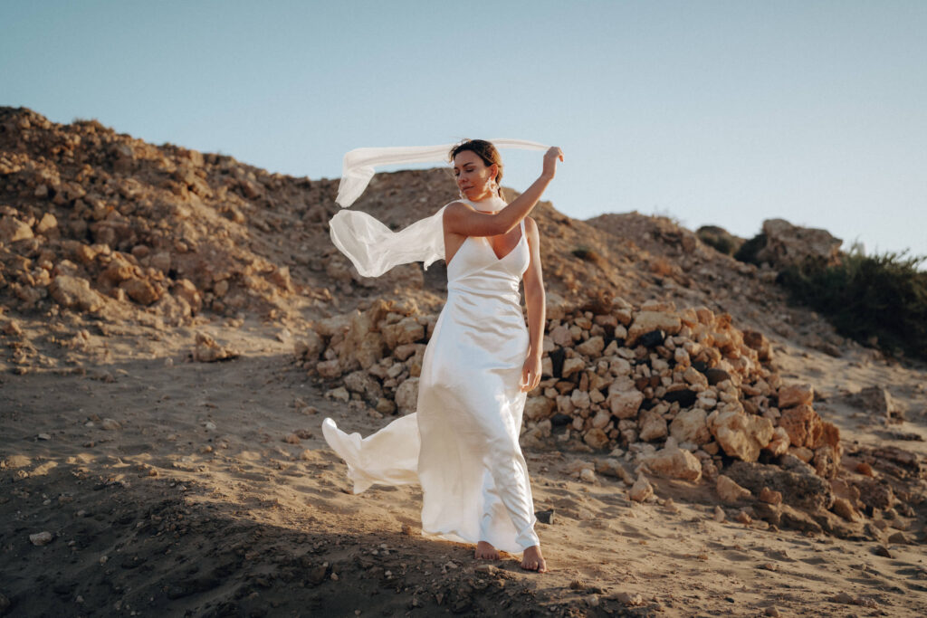Eine Frau im Brautkleid steht am Strand von Fuerteventura mit wehendem weißen Schal.