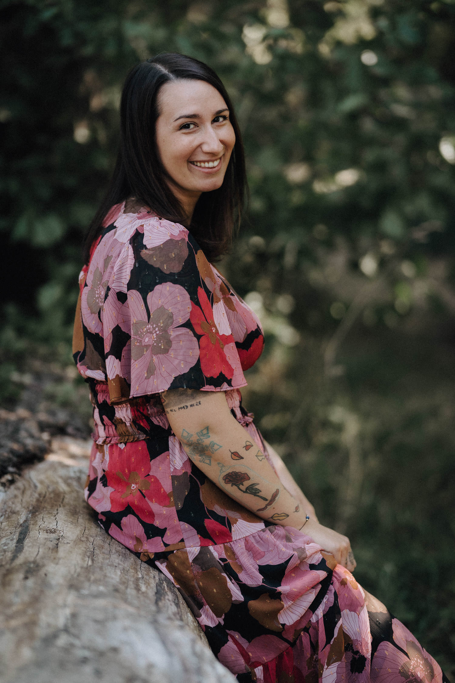 Eine Frau im rosa Blumenkleid lehnt an einem Baumstamm und schaut in die Kamera.